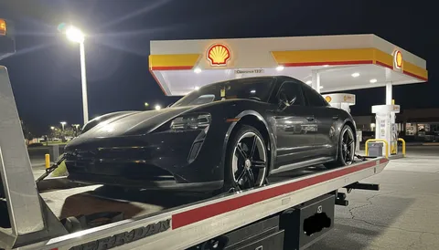 Black vehicle being towed on a flatbed tow truck with a Shell gas station visible in the background in Paramount, California.